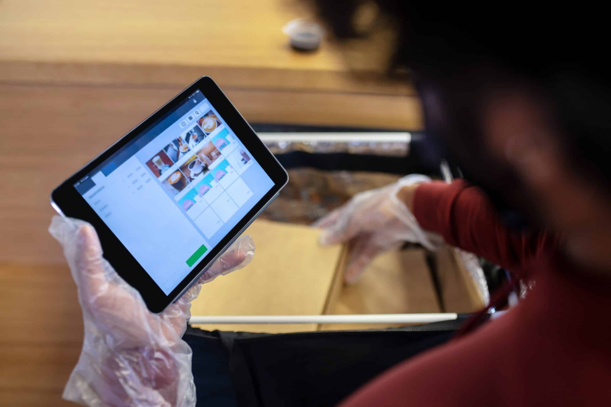 Person with gloves holding a tablet displaying a menu, packing items into a delivery bag.